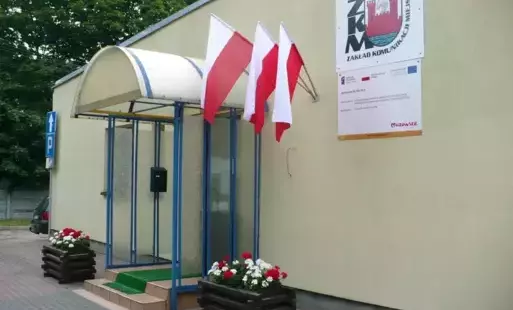 Zdjęcie przedstawia wejście do budynku Zakładu Komunikacji Miejskiej, budynek w kolorze żółtym, na ścianie logo ZKM, flagi polski zawieszone na ścianie budynku.