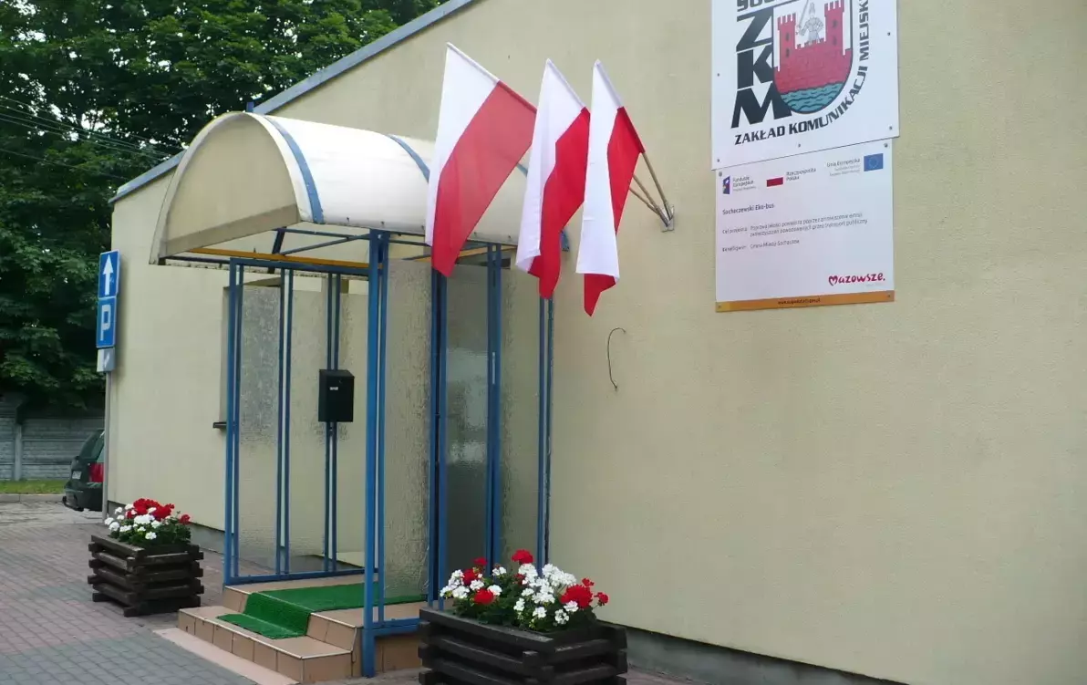 Zdjęcie przedstawia wejście do budynku Zakładu Komunikacji Miejskiej, budynek w kolorze żółtym, na ścianie logo ZKM, flagi polski zawieszone na ścianie budynku.